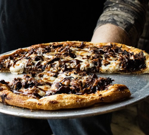 A person holding a large slice of pizza topped with melted cheese and dark, savory toppings on a metal tray.