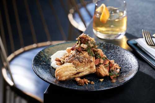 A flaky croissant topped with savory toppings, served on a dark plate alongside a glass of cocktail with a citrus garnish.