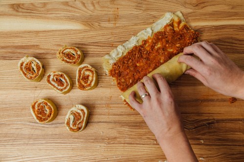 Hands rolling a pastry filled with meat sauce, with sliced pinwheels of the pastry arranged nearby on a wooden surface.