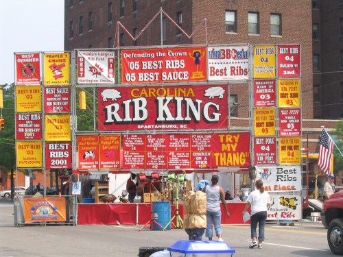 Colorful signage for "Carolina Rib King" in Spartanburg, SC, showcasing awards and menu items at a food festival.
