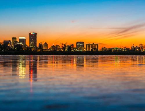 City skyline at sunset, reflecting on calm water with vibrant orange and blue hues in the sky.