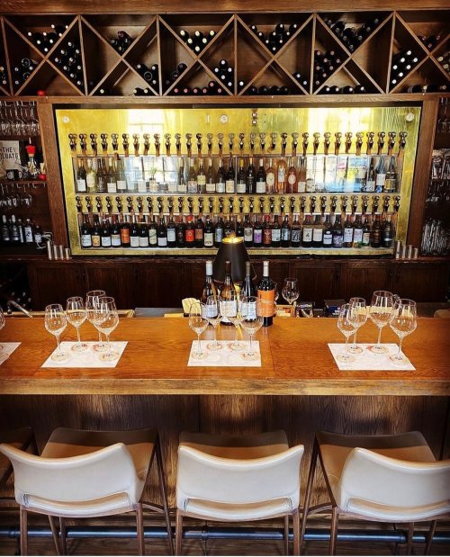 A stylish bar with a wooden counter, wine glasses, and a wall of wine bottles and taps in the background.