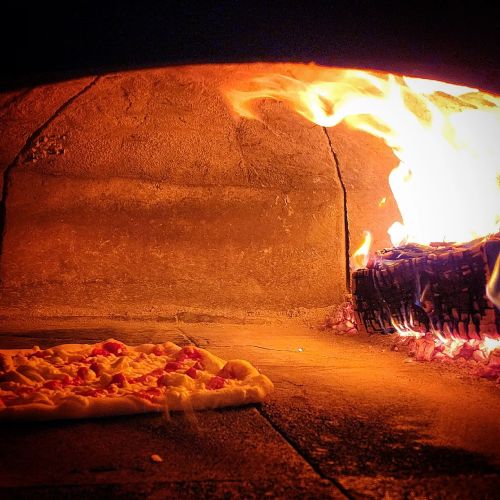 A pizza baking in a wood-fired oven, with flames and glowing embers visible in the background.