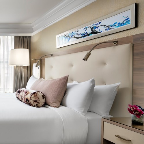 A stylish hotel bedroom featuring a plush bed with decorative pillows, a bedside lamp, and modern artwork on the wall.