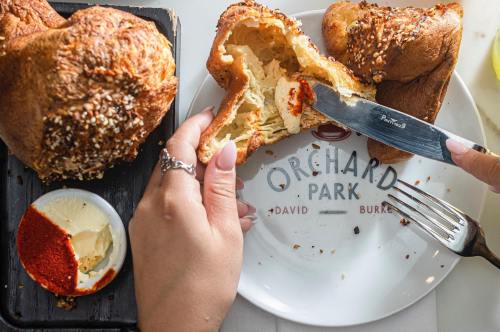 A hand holding a knife cutting into a warm, flaky pastry on a plate with butter and seasoning.
