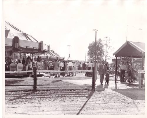 A busy outdoor fairground scene with crowds near attractions and tents, captured in black and white.
