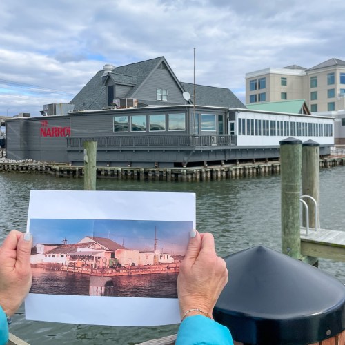 A person holds a photo of a waterfront building, comparing it to the current view of the same location.