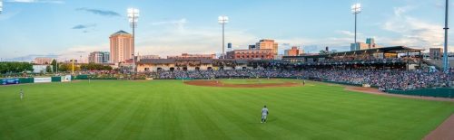 A panoramic view of a baseball stadium filled with fans, with city buildings and a sunset in the background.