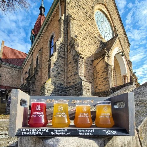 Four colorful drinks on a wooden tray in front of a historic stone building, with a clear blue sky above.