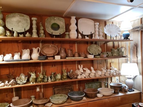 A wooden shelf displays various pottery pieces, including vases, bowls, and decorative bunnies, against a rustic backdrop.