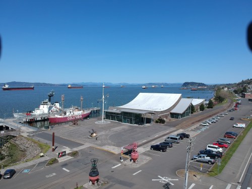 A view of a harbor with ships, a large building, and parked cars along the waterfront under a clear blue sky.