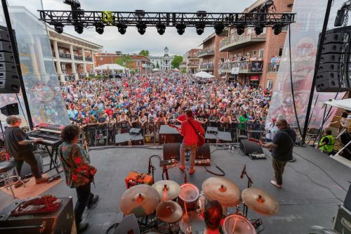 A lively outdoor concert scene with a large crowd facing the stage, musicians performing, and a vibrant atmosphere.