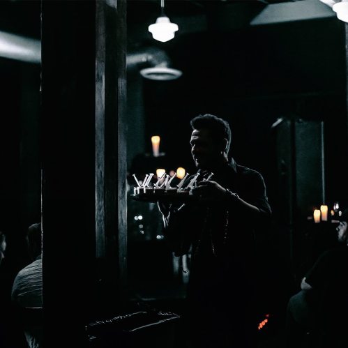 A silhouette of a person holding a tray with candles, surrounded by dim lighting and a crowd in the background.