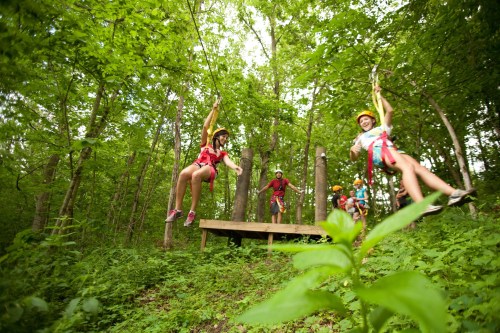 Two children on zip lines in a lush green forest, with adults and trees in the background.