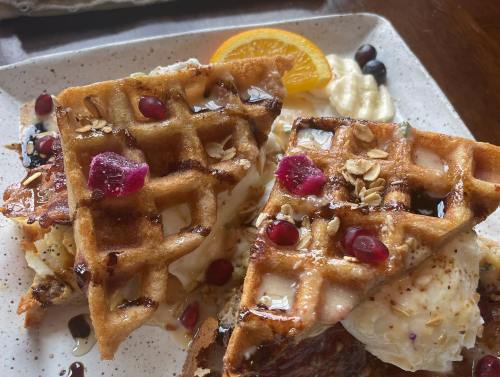 Two golden waffles topped with cream, berries, and a drizzle of syrup, garnished with orange and pomegranate seeds.