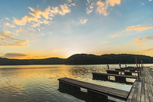 A serene lake at sunset, with wooden docks and silhouettes of people fishing against a colorful sky.