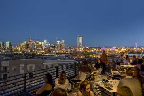 A rooftop bar scene at dusk, with people socializing and a vibrant city skyline illuminated in the background.