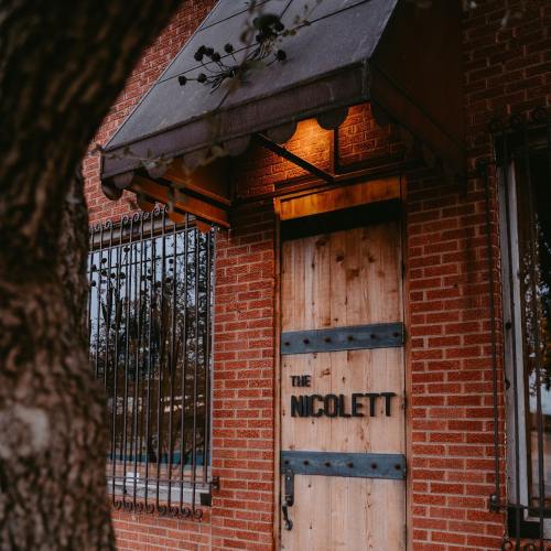 A rustic wooden door with "The Nicolette" sign, framed by a brick wall and a canopy, partially obscured by a tree.