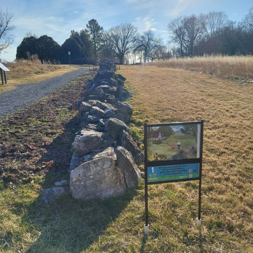 A rocky path alongside a grassy area, with an informational sign depicting a scenic view. Trees in the background.