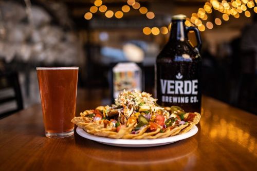 A plate of nachos topped with vegetables and a drink beside a growler from Verde Brewing Co., set in a cozy restaurant.