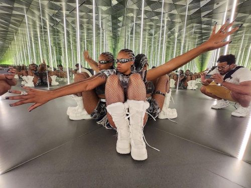 A person in a mirrored room poses with outstretched arms, wearing sunglasses, fishnet stockings, and white boots.
