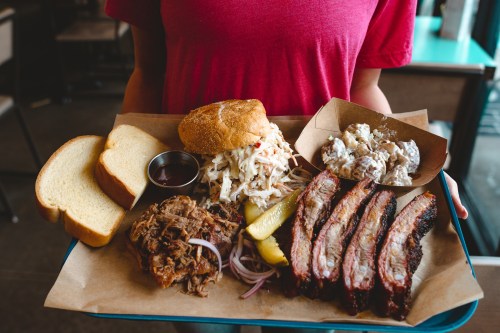 A person holding a tray with pulled pork, ribs, coleslaw, potato salad, a sandwich, and slices of bread.