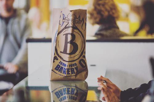 A brown paper bag with a large "B" logo sits on a table, reflecting in the glass surface, with people in the background.