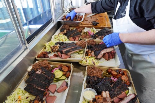 A person in gloves arranges trays of barbecue with meats, sides, and pickles in a food service setting.