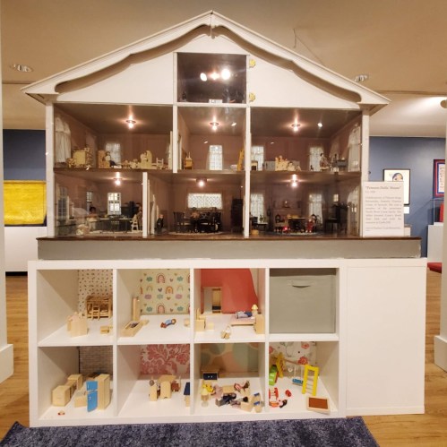 A detailed dollhouse display with multiple rooms, showcasing miniature furniture and toys in a museum setting.