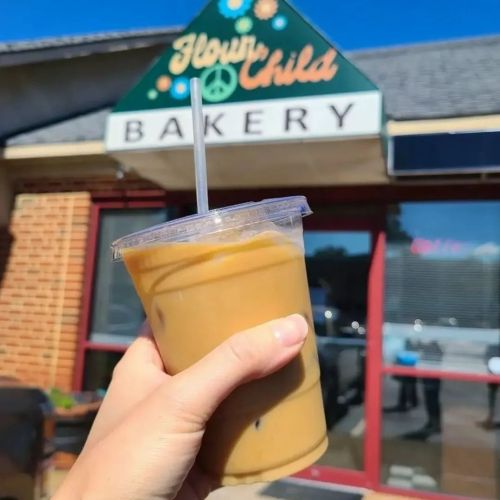 A hand holding an iced drink in front of a bakery with a colorful sign that reads "Flour Child Bakery."