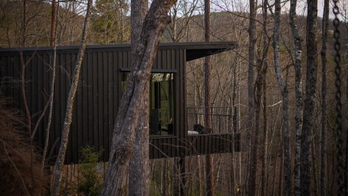 A modern black cabin perched among trees in a forest, featuring large windows and a small balcony.