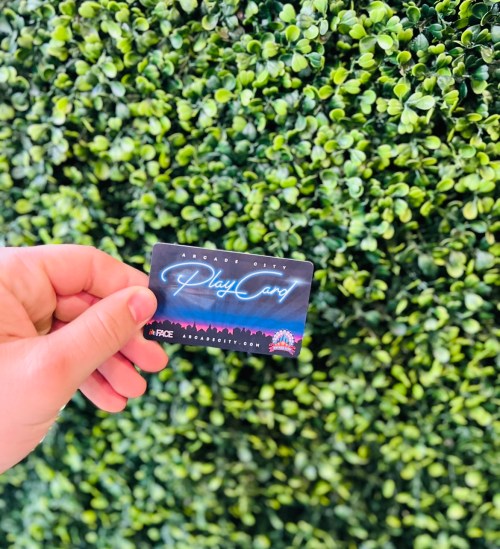 A hand holding a "Play Card" in front of a green, leafy background.