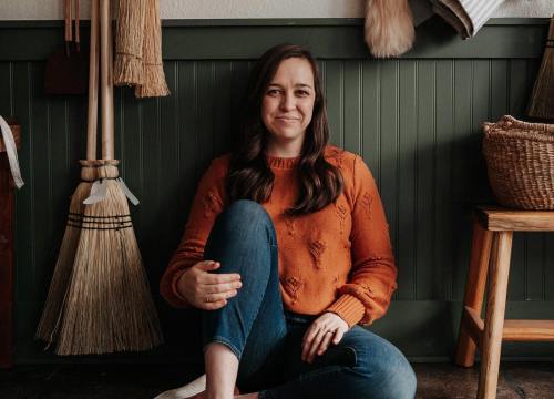 A woman in an orange sweater sits on the floor, smiling, surrounded by brooms and a woven basket.