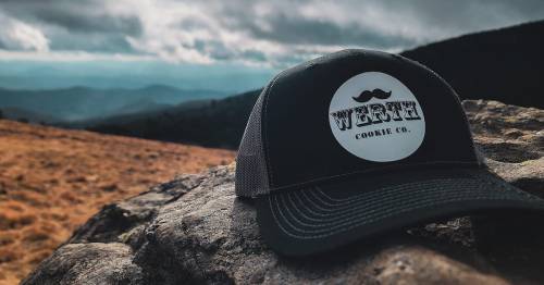 A black cap with a circular logo reading "WERT COOKIE CO." rests on a rock, with a scenic mountain backdrop.