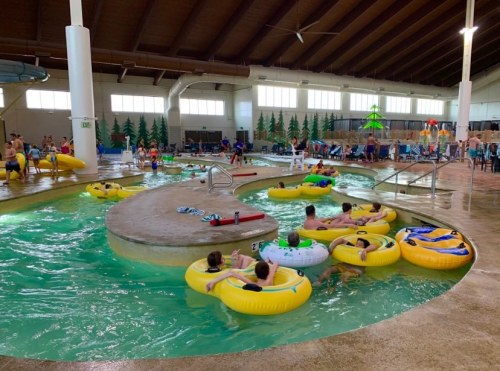 Indoor water park scene with people floating on yellow inner tubes in a lazy river, surrounded by trees and play areas.
