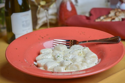 A plate of creamy gnocchi topped with herbs, accompanied by a fork, with a bottle of wine in the background.