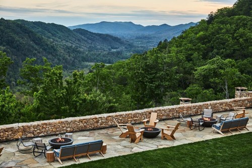 A scenic view of mountains and valleys from a patio with fire pits and seating, surrounded by lush greenery.