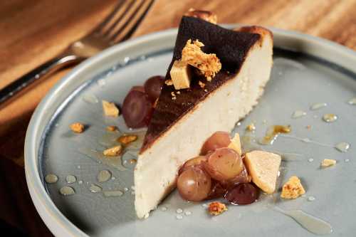 A slice of cheesecake topped with grapes and honeycomb on a blue plate, with a fork in the background.