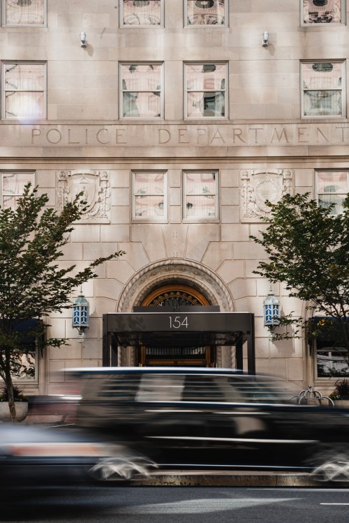 Facade of a police department building with a large entrance and trees, while a car passes by in motion.