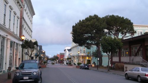 Quiet street scene with trees, historic buildings, and parked cars under a cloudy sky.