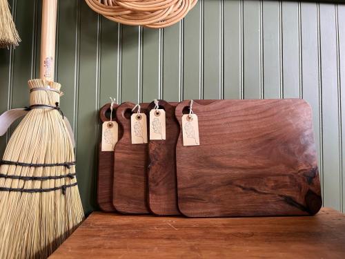 A set of wooden cutting boards with tags, displayed alongside a straw broom against a green wall.