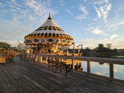 A colorful carousel on a wooden pier, reflecting in calm water under a blue sky with wispy clouds.