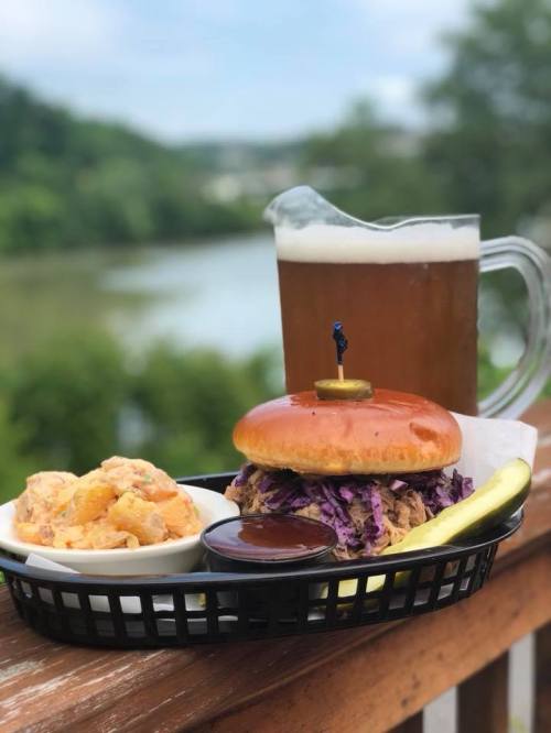 A pulled pork sandwich with coleslaw, a pickle, and a side of potato salad, alongside a pitcher of beer by a river.