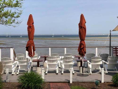A seaside deck with white chairs and red umbrellas, overlooking a calm beach and distant sailboats on the water.