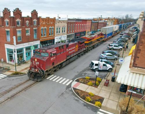 A red train passes through a small town street lined with shops and parked cars on a cloudy day.