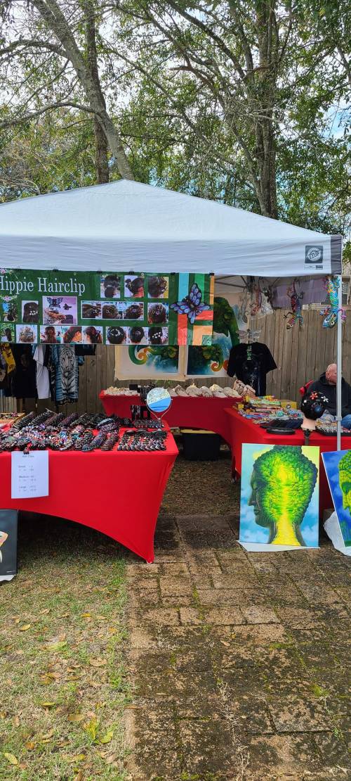 A vibrant market stall with red tables displaying hair clips, art, and crafts under a white tent surrounded by trees.