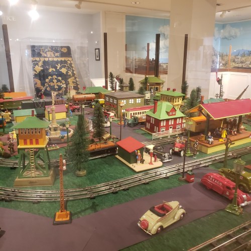A colorful miniature train set with buildings, trees, and figures, displayed in a museum setting.