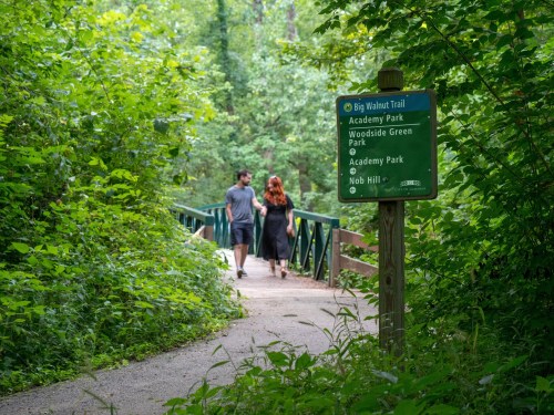A couple walks along a path in a lush green area, with a trail sign visible nearby.