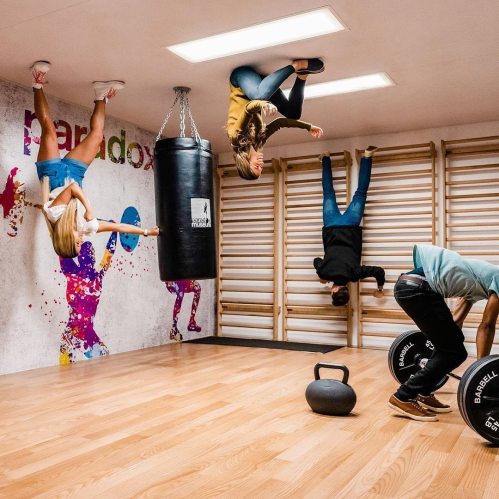 A gym with people exercising in various positions, including upside down, against a colorful wall and fitness equipment.