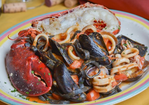 A colorful plate of seafood pasta featuring lobster, squid, mussels, and black noodles in a rich tomato sauce.